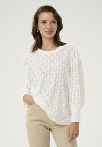 Pull en maille blanche avec un motif en losange, col rond et manches longues bouffantes. Associé à un pantalon beige.