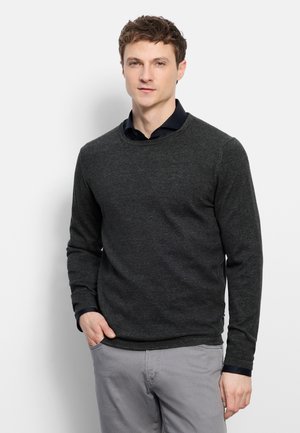 Jonge man staat met hand in zak, draagt een donkergrijze trui over een zwart overhemd met kraag en lichtgrijze broek tegen een witte achtergrond.