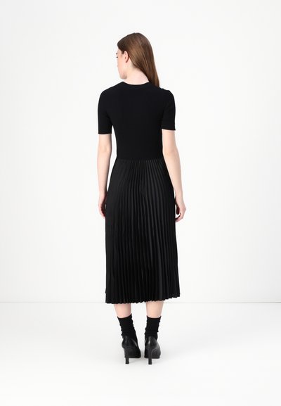Robe noire avec un haut ajusté à manches courtes et une jupe midi plissée. Texture lisse sur le haut, plis fluides en bas, portée avec des chaussettes à mi-mollet et des talons.