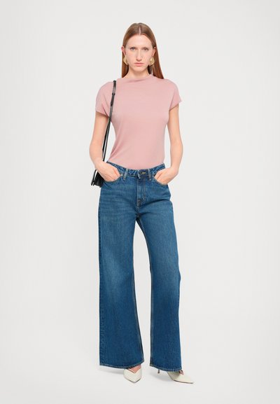 Un haut rose clair à manches courtes associé à un jean en denim bleu à jambes larges. Les détails notables incluent une taille haute, des coutures contrastées et des chaussures à bout pointu crème.