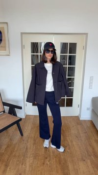 Žena s černou čepicí s červeným srdcem, slunečními brýlemi, černou bundou, bílým tričkem, černými kalhotami a bílými teniskami stojí uvnitř na dřevěné podlaze.