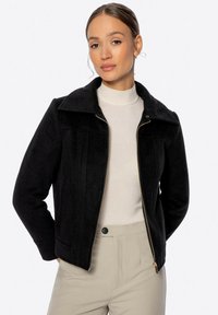 Chaqueta de mezcla de lana negra con cremallera frontal, gran cuello y bolsillos laterales. Presenta un corte entallado, superficie texturada y líneas limpias.