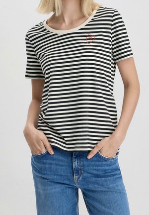 Kvinde iført kortærmet sort-hvid stribet T-shirt med lille rødt hjerte nær kraven, hænderne i lommerne på blå jeans.