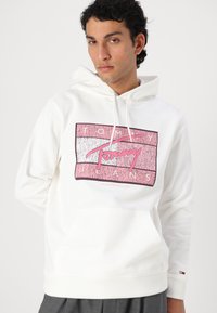 Hvit hettegenser med et rosa og sølv grafisk logo, justerbar snøring på hetten og en frontlomme. Ribbestrikkede mansjetter og bunn.