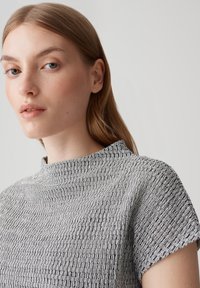 Haut texturé gris avec un col montant et des manches courtes, arborant un motif répétitif de petites boucles entrelacées.