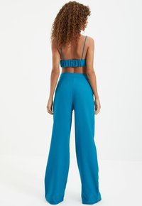 Tyrkysový dvoudílný outfit, skládající se z crop topu s volánkovým zadním dílem a tenkými ramínky, doplněný o kalhoty s širokými nohavicemi, oba vyrobené z lehkého materiálu.