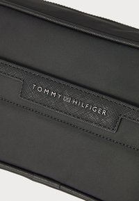 Pochette in tessuto nera con etichetta nera testurizzata e testo bianco in rilievo "TOMMY HILFIGER", con dettagli di cucitura discreti lungo i bordi.