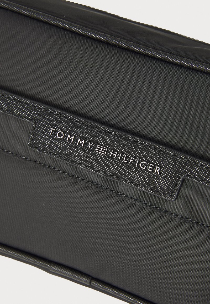 Pochette en textile noire avec une étiquette texturée noire portant le texte en relief "TOMMY HILFIGER" et des détails de couture subtils le long des bords.