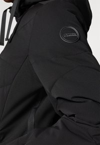 Melnā polsterētā jaka ar teksturētu virsmu, kurai uz piedurknes ir apļveida logo. Tajā ir kapuce un regulējami aukliņas.