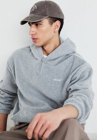 Felpa grigia in pile con cappuccio e lacci, dotata di un logo bianco di Levi's. Indossata con un cappello marrone, presenta un look casual e stratificato.