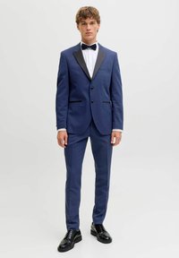 Jauns vīrietis stāv, tērpies zilā smokings ar melniem atloki, baltu krekliņu, melnu tauriņu un melnus oficiālus apavus uz vienkārša gaiša fona.