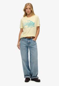 Gul bomulds t-shirt med blå tekst, parret med lyseblå denimjeans og brune sko. Modellen står med hænderne på hoften foran en hvid baggrund.