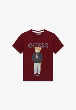 T-shirt bordeaux avec un ours en peluche portant un sweat à capuche noir et un pantalon clair, nom de la marque "GUESS" au-dessus, texte "Est. 1981" en dessous.
