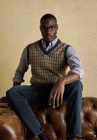 Tumedate V-kaelusega, mustriga sabakiri, pehme kampsun, tumesinises ja beežis, sinise ja valge triibulise pluusi peal. Tumedad teksad ja pruun nahkdiivan.