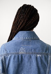 Veste en denim bleu clair avec un col, présentant des détails brodés subtils dans le dos et un patch au-dessus du dos.