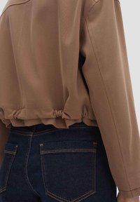 Korte bruine jas met een geraffineerde zoom, gemaakt van een gladde stof. Gecombineerd met donkerblauwe jeans met twee achterzakken en oranje stiksels.