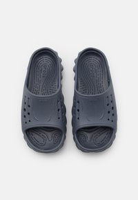 Marinblå slip-on skor gjorda av mjuk, strukturerad gummi med dräneringshål och ett vågigt souldesign för grepp. Ren design och rundade kanter.