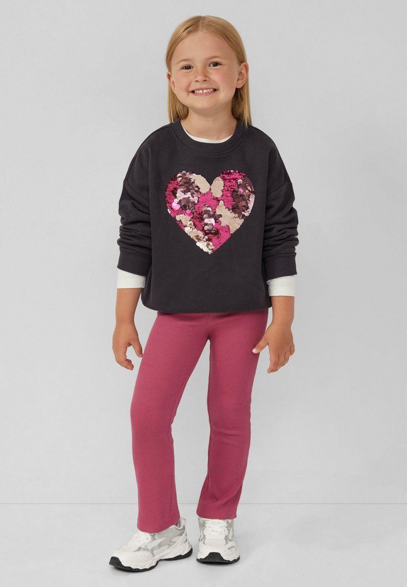 Dunkelgrauer Pullover mit einem Herzdessin aus mehrfarbigen Pailletten, kombiniert mit rosa gerippten Leggings und weißen Turnschuhen.