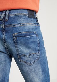 Blå denimjeans med en avslappet passform, med en teksturert overflate, oransje sømmer og en firkantet baklomme med et subtilt design.