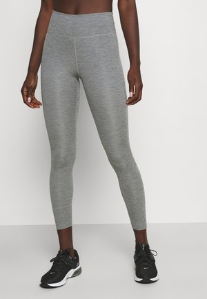 Leggings gris avec une texture lisse, une taille haute et un design ajusté. Associés à des chaussures de sport noires et un fond uni.