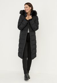 Zwarte pufferjas met een lang model, gevoerde capuchon met bont, doorgestikte textuur en een ritssluiting aan de voorkant. Draagt deze over een zwarte broek en enkellaarzen.