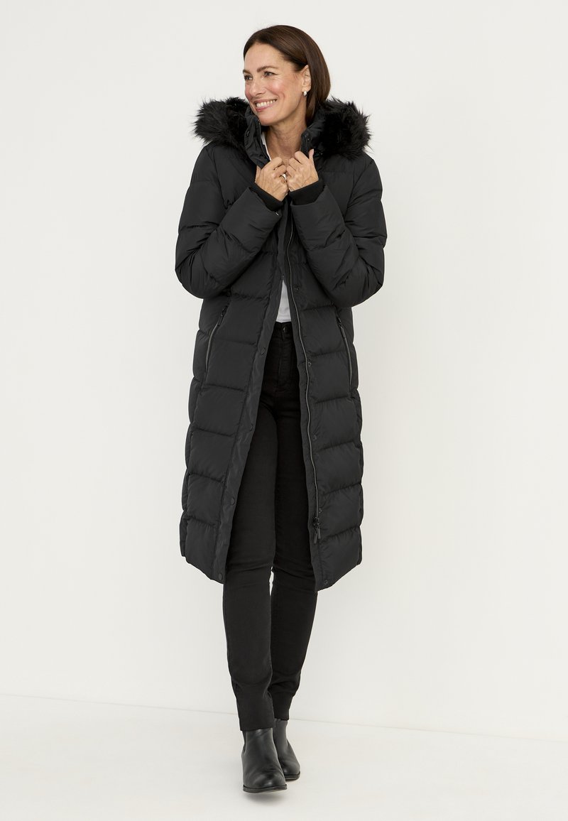 Zwarte pufferjas met een lang model, gevoerde capuchon met bont, doorgestikte textuur en een ritssluiting aan de voorkant. Draagt deze over een zwarte broek en enkellaarzen.