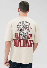 T-shirt beige avec un graphique d'une main de squelette tenant des cartes à jouer et la phrase "TOUT OU RIEN" en lettres rouges en gras.