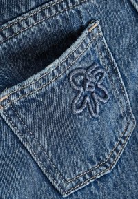 Un bolsillo de vaqueros de mezclilla azul oscuro con un detalle de flor bordada en un tono azul a juego. La tela muestra una trama texturizada y un ligero desgastado.