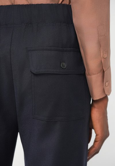 Navy trousers with a textured fabric, featuring an elastic waistband and a side pocket with a black button. Brown shirt sleeve is visible.