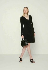 Mulher em vestido envolvente preto de manga longa, segurando uma bolsa preta, usando sandálias de salto pretas e colares de ouro em camadas, em pé sobre um fundo claro.