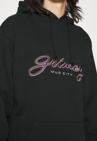 Černá mikina s kapucí, která má na sobě vyšívaný text "Grimey" a "Mud City" v purpurové a zlaté. Měkký materiál s velkou přední kapsou.
