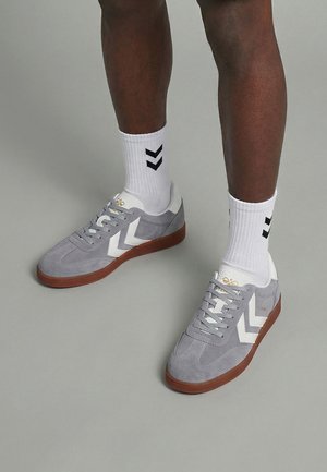 Person iført grå ruskindsneakers med hvide chevron-striber, hvide sokker med sorte chevron-symboler, stående på en grå gulv.