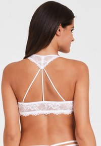 Weißes Spitzen-Bralette mit hohem Halsausschnitt, dünnen Trägern und floralem Muster am Bund. Der Rücken hat ein T-Träger-Design.