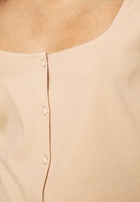 Beige ribbad topp med rundad halsringning och små knappar fram, som framhäver det texturerade tyget och den minimalistiska designen.