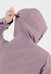 Violetti vedenpitävä takki, jossa on kiristettävä huppu. Ominaisuuksia ovat sileä kangas, näkyvät saumat ja takana oleva säätölenkki.