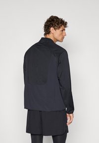 Mænds sort jakke med en struktureret bagpanel, der har høj krave og lange ærmer. Materialet er let og vandafvisende.