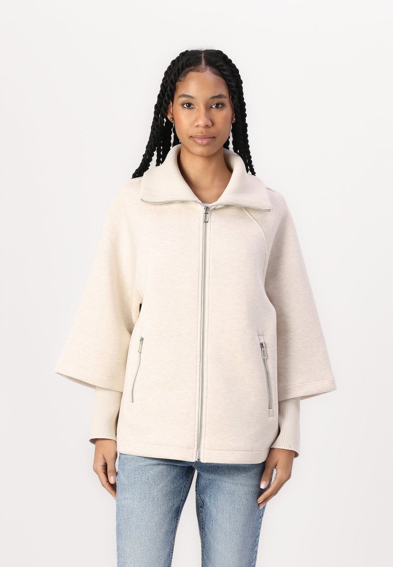 Beige, overdimensioneret zip-jakke med højt krave, ribbede manchetter og sidelommer med lynlås. Blød tekstur og brede ærmer giver komfort.