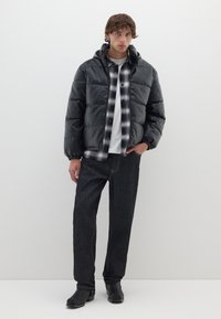 Chaqueta de plumón negra con capucha, material brillante, puños elásticos, superpuesta a una camisa de cuadros, combinada con jeans oscuros y zapatos negros.