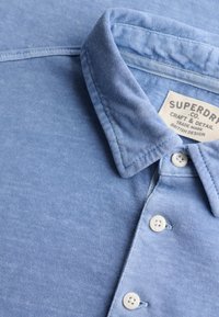 Light blue button-up shirt collar and buttons with a visible tag reading "Superdry Co. Craft & Detail British Design."