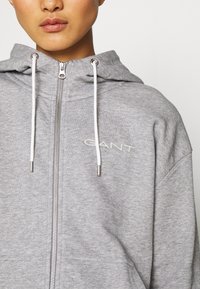 Felpa grigia con zip, realizzata in tessuto morbido, con cerniera frontale, coulisse bianche e il nome del marchio "GANT" ricamato in bianco.