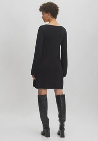 Schwarzes Strickkleid mit langen Ärmeln, ausgestattet mit einem gewellten Ausschnitt und Saum. Kombiniert mit hohen schwarzen Stiefeln, die eine schlanke Silhouette betonen.