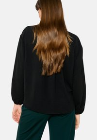 Black long-sleeve top with a relaxed fit, rounded neckline, and balloon sleeves, made from a soft texture. Hair falls over the back.