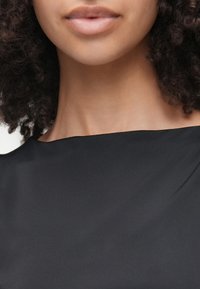 Μαύρη, λεία υφασμάτινη μπλούζα με φαρδύ λαιμόκομμα, που αποκαλύπτει τον λαιμό και τα χείλη, με φυσικά, ουδέτερα μακιγιάζ και απαλά μπούκλες.