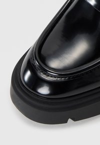 Μαύρο παπούτσι από πατενταρισμένο δέρμα με κομψό σχέδιο, στρογγυλή μύτη και παχύ, υφασμένο καουτσούκ σόλα. Έχει διακριτική ραφή κατά μήκος της άκρης.