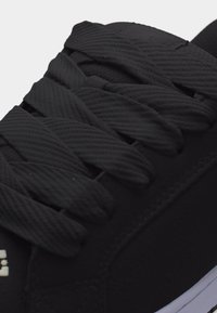 Κοντινή λήψη μαύρων κορδονιών με υφή υφάσματος πλεγμένων μέσα από οπές σε μαύρο αθλητικό παπούτσι, με ορατά άκρα λευκής σόλας.