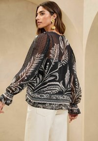 Bluză neagră transparentă cu modele albe florale și paisley, cu manșete lungi și bufante, și cu manșete elastice. Purtați peste pantaloni off-white.