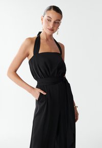 Woman with slicked-back hair wearing a black halter jumpsuit with a waist tie, one hand in pocket, and gold earrings and bracelet.