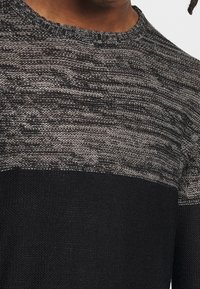 Sweter dziergany z fakturowanym szarym wzorem na górze, jednolity czarny dół oraz okrągły dekolt. Materiał wydaje się miękki i ciepły.