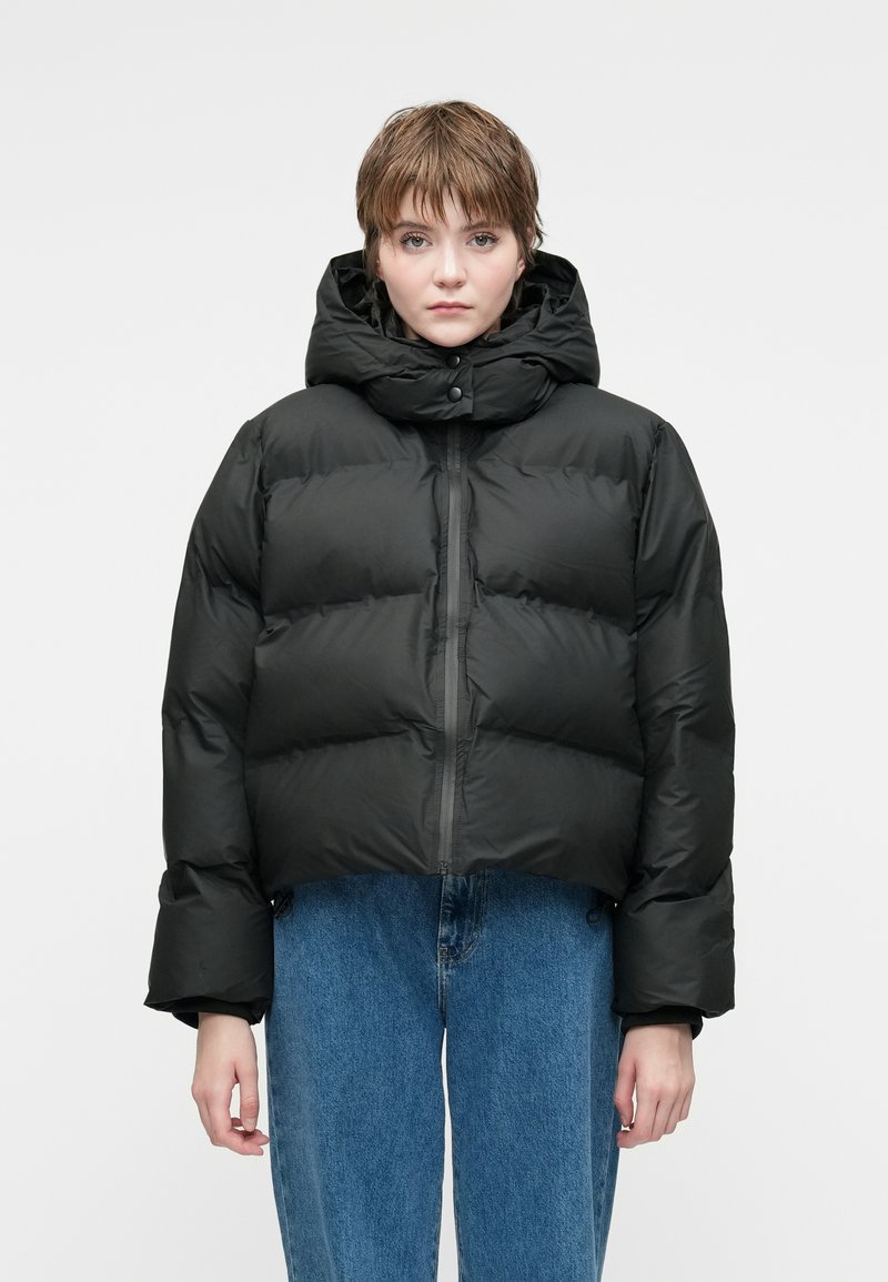 Geacă puffer neagră, scurtă, cu guler înalt, închidere cu capse și textură netedă. Asortată cu blugi denim deschis la culoare.