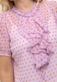 Blusa lila con pequeños patrones geométricos naranjas, con pliegues en la parte frontal, mangas cortas abullonadas y cierre de botones en la parte delantera. Tela suave y ligera.